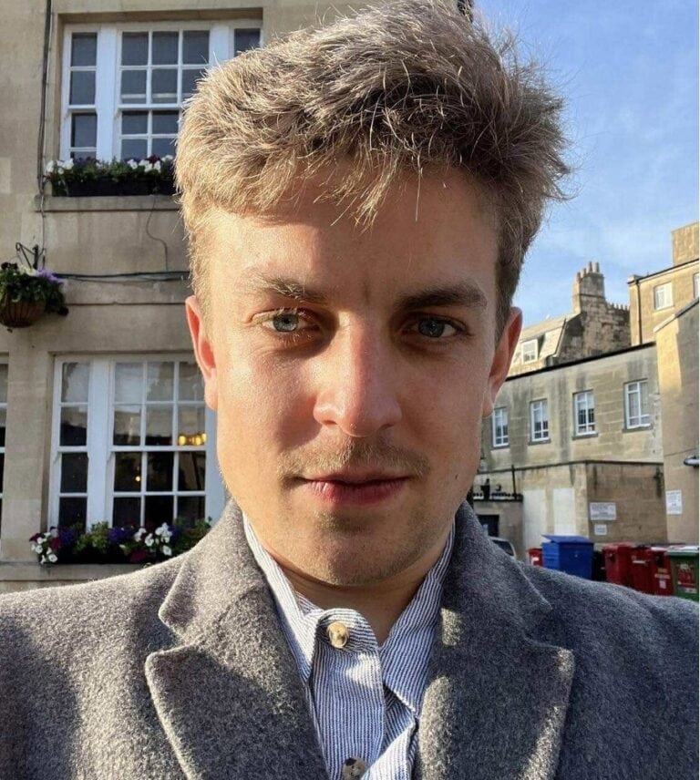 Portrait of a young man with light brown hair and blue eyes, wearing a grey coat and a striped shirt, standing outdoors in East London on a sunny day.